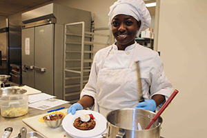 woman in chef's uniform holds up a dish she prepared